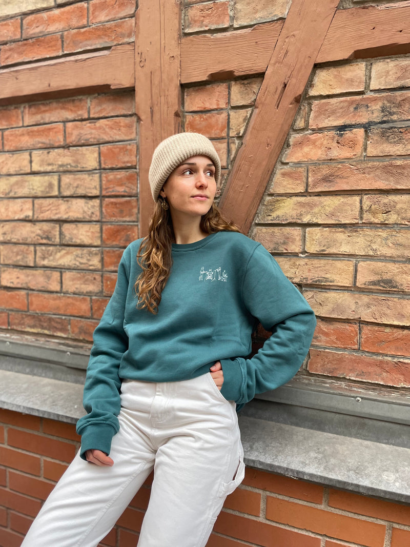 Frau lässig an Wand gelehnt in nachhaltigem Sweatshirt in der Farbe Mediterranea Green. mit gezeichnetem Cat Band-Motiv (Katzenband mit Keyboard, Schlagzeug, Mikrofon, Gitarre). Druck-Motiv sitzt über der linken Brust, Foto von Ungerade Wien
