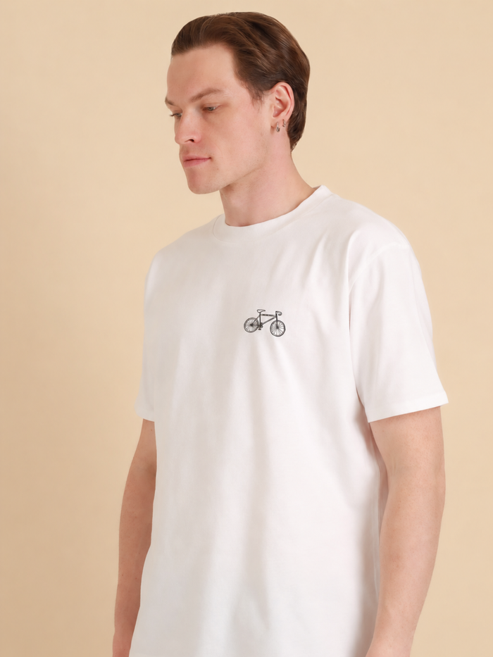 Auf dem Foto: ein Mann, fotografiert von der schrägen Front-Ansicht. Er trägt ein weißes Bio-Baumwolle Shirt (GOTS) mit Fahrrad/Bike-Druck-Motiv über der linken Brust. Es handelt sich um ein Oversized Unisex Shirt. Der Druck des Motivs ist schwarz. Das Bike ähnelt einem Mountainbike. Produktfoto von Ungerade Wien