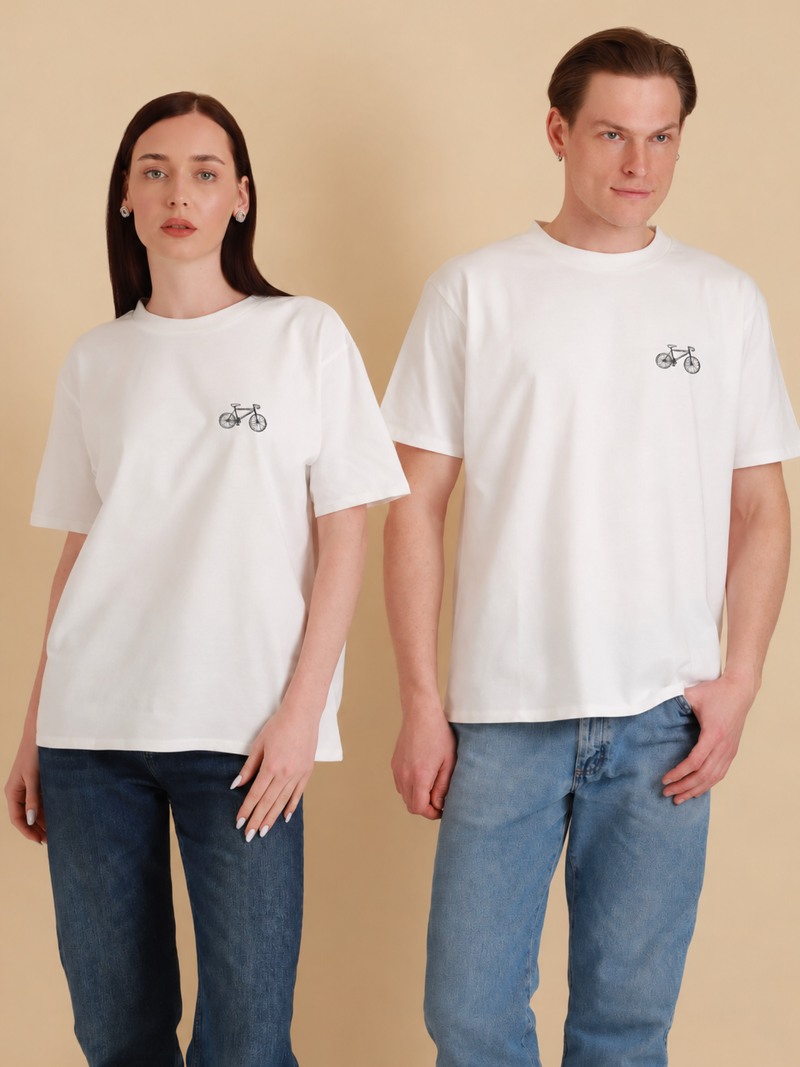 Auf dem Foto: Eine Frau (Links) und ein Mann (rechts), jeweils mit dem gleichen weißen Bio-Baumwolle Shirt (GOTS) mit Fahrrad/Bike-Druck-Motiv über der linken Brust. Es handelt sich um ein Oversized Unisex Shirt. Der Druck des Motivs ist schwarz. Das Bike ähnelt einem Mountainbike. Produktfoto von Ungerade Wien