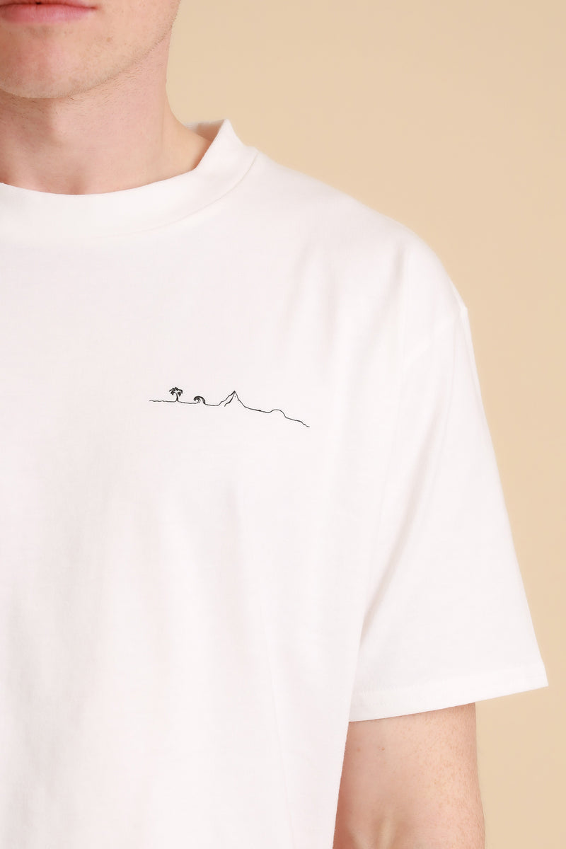 Insel-T-Shirt Men: auf dem Foto ist ein Mann zu sehen. er trägt ein weißes T-Shirt mit schwarzem Druck. zu sehen ist  ,von links nach rechts durchgehend verbunden, eine Palme, Welle, ein Berg, eine aufgehende Sonne. Alle Motive zusammen sollen eine Insel symbolisieren. Produktfoto von Ungerade Wien