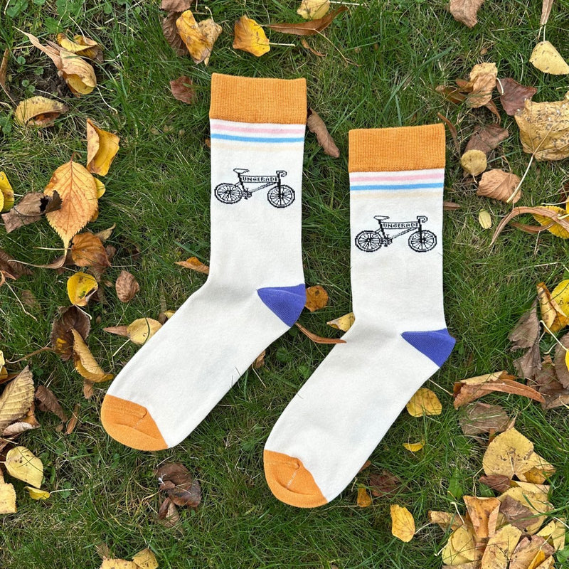 Bike Classic – Bio-Baumwollsocken mit sportlichem Design .Die Socken liegen im grünen Gras, umgeben von Herbstblättern. Der Sockenbund ist ca. 4 cm breit, ocker-gelb. Darunter befinden sich 3 Querstreifen (ca 3-5mm dick) in rosa, hellblau, hellgelb/beige. Darunter liegt das gezeichnete Bike-Motiv. in der Querstange des Bikes steht "Ungerade". Die Ferse ist dunkellila und die Sockenspitze ockergelb. - Ungerade Wien