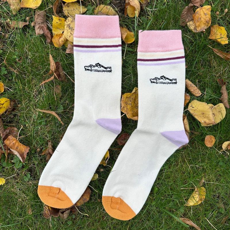 Lazy Sandals – Bio-Baumwollsocken mit Sandalen-Feeling. das Motiv zeigt einen gezeichneten Schlappen/Hausschuh. Der Sockensaum ist ca. 4cm breit und hellrosa. darunter liegen 3 Querstreifen (ca. 3-5mm dick), hellbeige/gelb, dunkelrot, Iila. 2cm darunter liegt das Druckmotiv. Die Sockenferse ist lila. Die Sockenspitze ist Ocker-gelb. Das Sockenpaar liegt im Gras, umgeben von Herbstblättern. Foto von Ungerade Wien